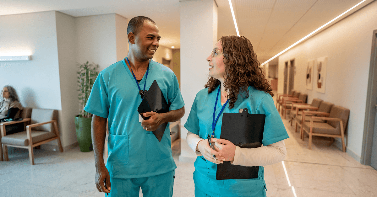 Two nurses in scrubs talking in a hospital corridor with clipboards