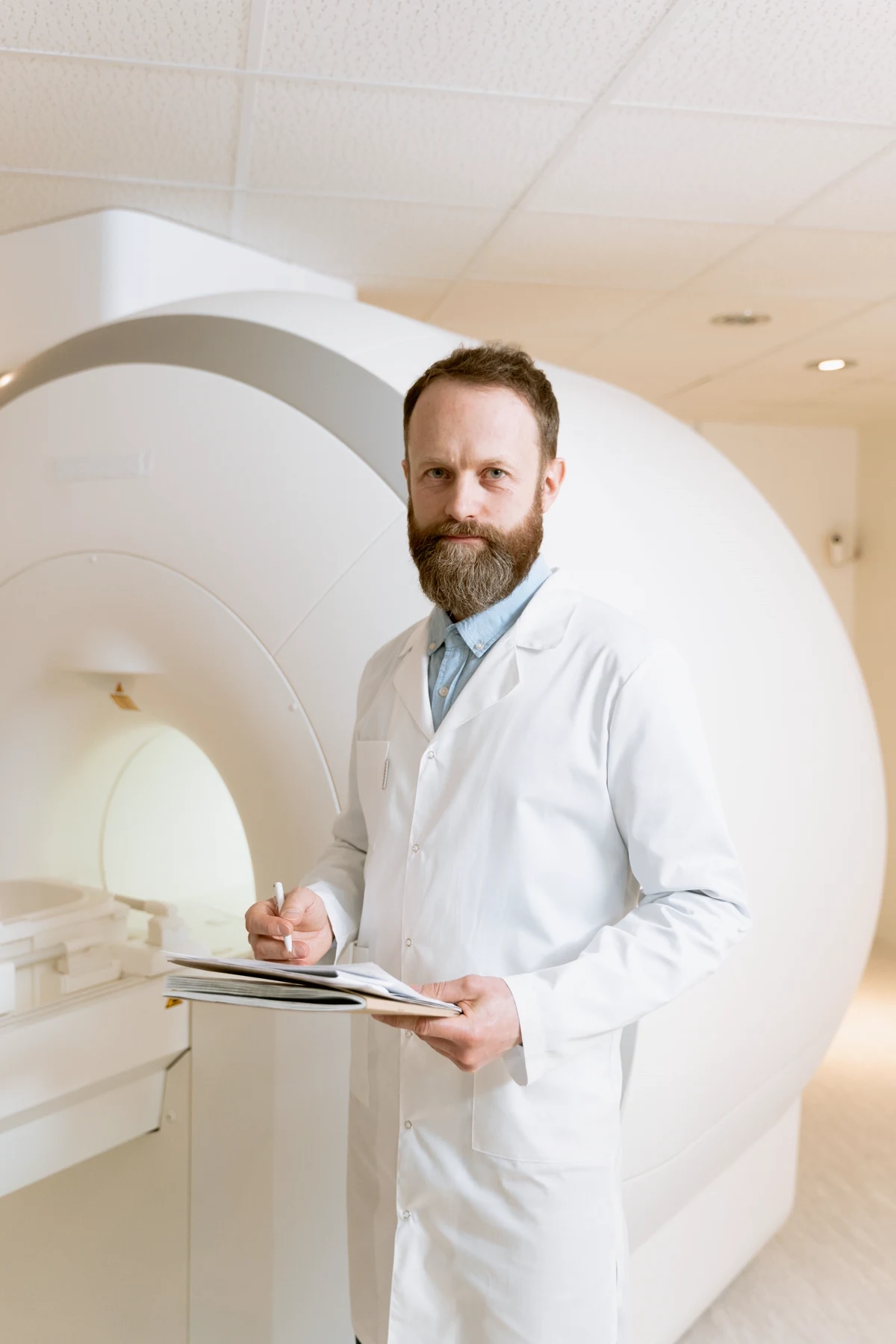 A bearded medical professional in a white coat holding a clipboard beside an MRI scanner.